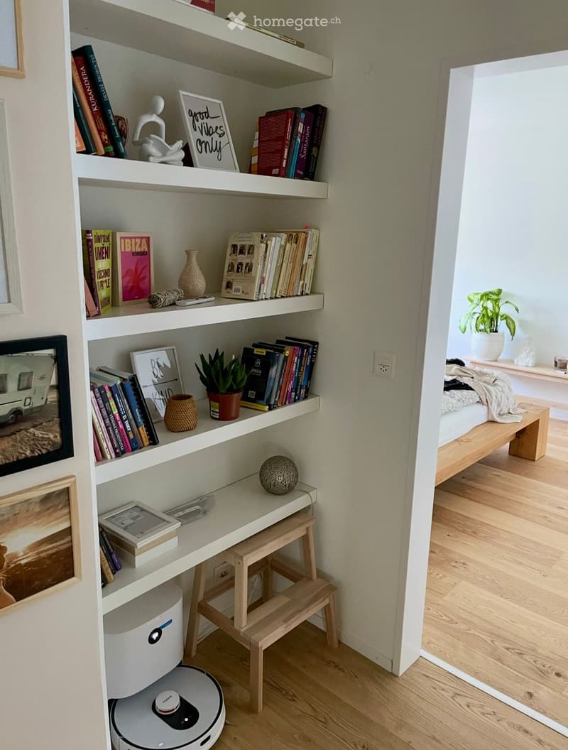 A cozy corner with white floating shelves filled with books, decorative items, and a small plant. A wooden bench and a small table with a stool are visible, along with a robot vacuum on the floor.