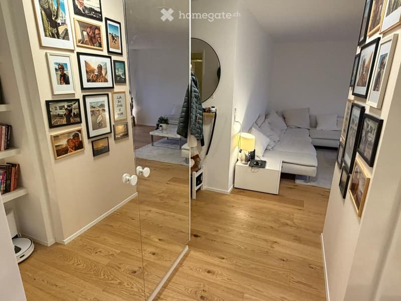 A compact, well-lit living space with a sofa, coffee table, nightstand, lamp, and framed pictures on the walls. The room features wooden flooring and a small entryway with a mirror.