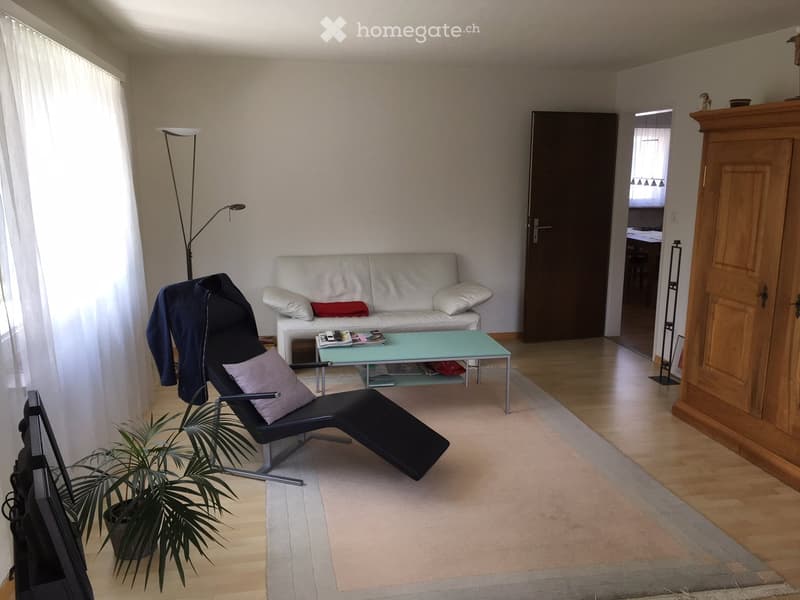 Modern living room with a white sofa, glass coffee table, black lounge chair, floor lamp, and a potted plant. The room features light-colored walls, wooden flooring, and a large window with white curtains.