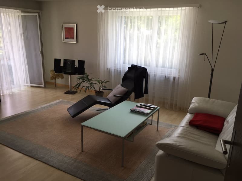 Modern living room with a light-colored sofa, a glass coffee table, a black lounge chair, a potted plant, a speaker system, and a floor lamp. The room features large windows with sheer curtains and a wooden floor.