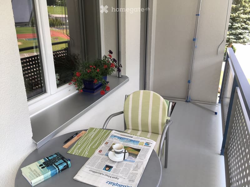 Balcony with a chair, small table, cup, book, newspaper, and flower pot. Offers a view of a sports field and trees.