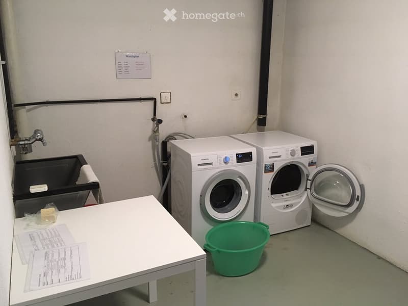 A small utility room with a washing machine, dryer, table, sink, and bucket.