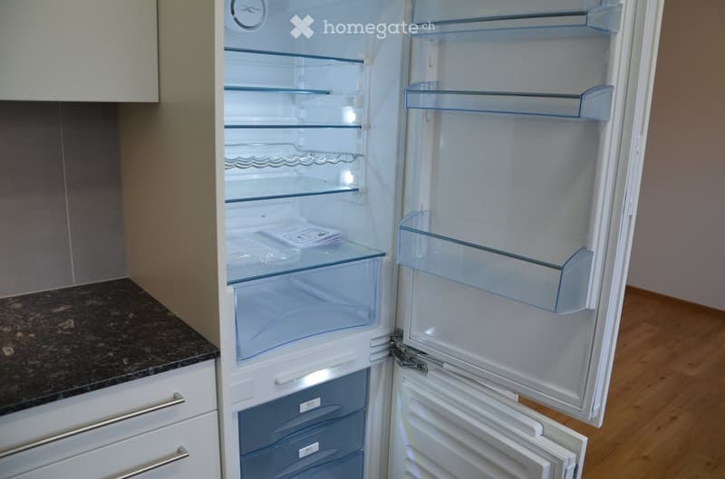 A modern kitchen with a white refrigerator featuring multiple glass shelves and compartments, including a freezer drawer. The refrigerator is open, revealing its interior with clear glass shelves and a few items inside.