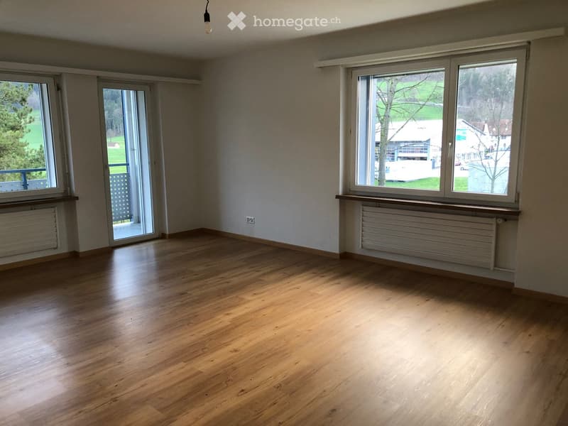 Empty room with wooden flooring, large windows, radiators, and a sliding glass door leading to a balcony.