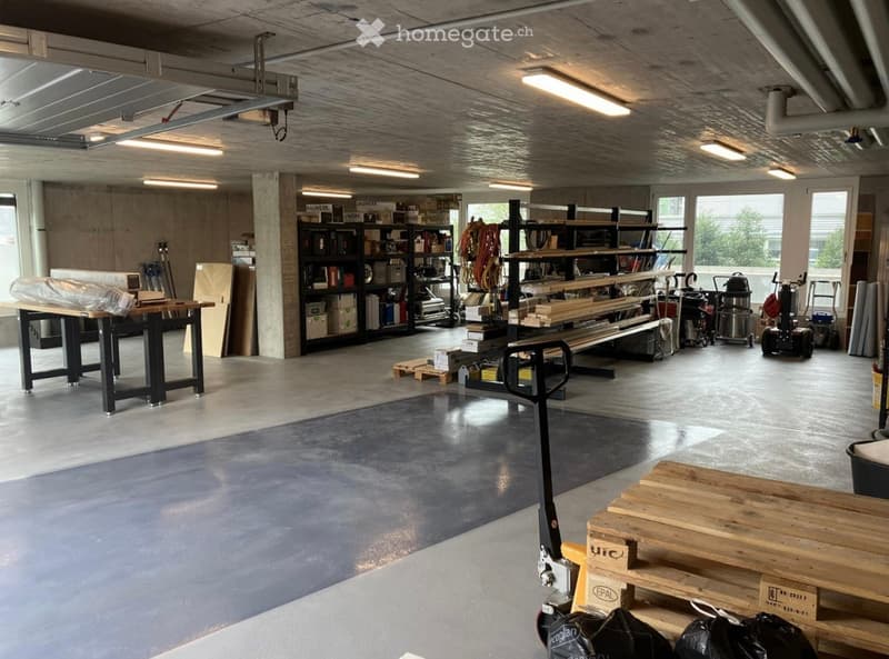A spacious workshop with concrete floors, exposed ceiling beams, and large windows. The room features a large workbench, metal shelving units filled with tools and supplies, and several pallets and machinery.