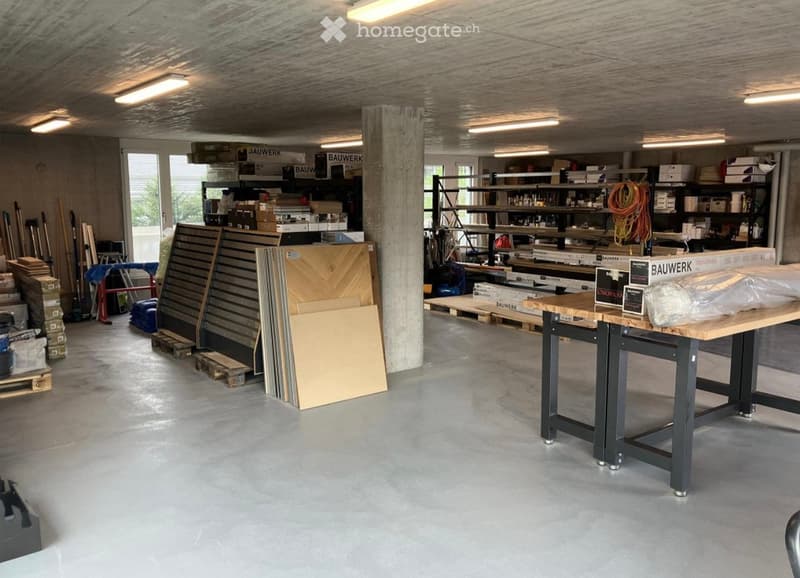 A spacious workshop with concrete floors, exposed wooden ceiling beams, and industrial-style lighting. Shelves hold tools and materials, and a large workbench is positioned in the foreground.