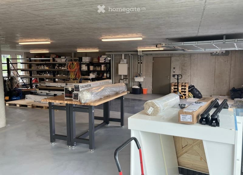Workshop or storage area with concrete walls, wooden tables, shelves filled with tools and materials, a sink, and a door.