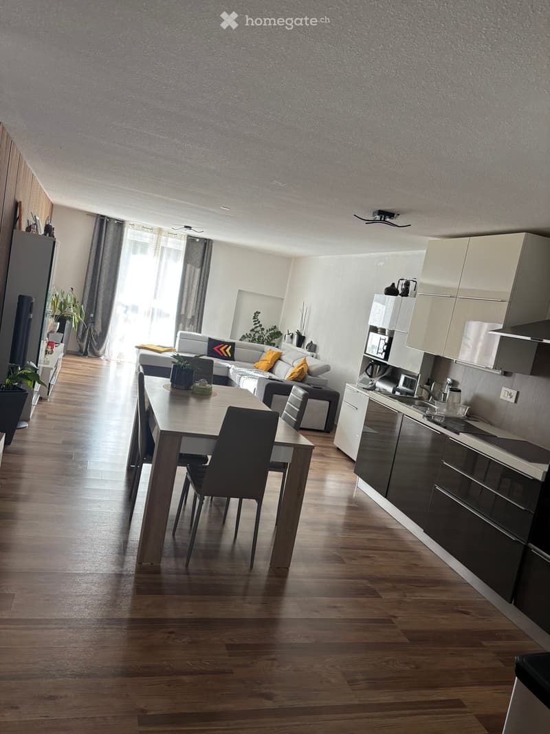 Modern open-plan living and dining area with a grey sectional sofa, wooden dining table, chairs, and a sleek kitchen with cabinets, appliances, and a sink.