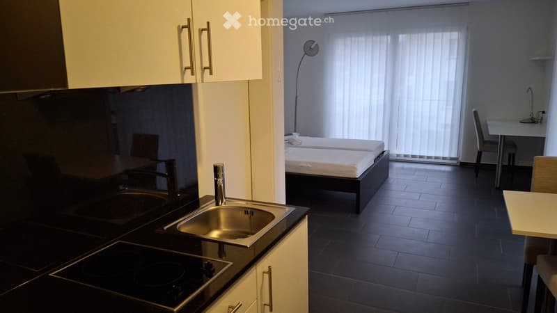 Modern, compact studio apartment with a kitchenette area featuring a sink, stove, and cabinets, and a sleeping area with a bed and chair. The room includes a desk and chair near a large window with blinds.