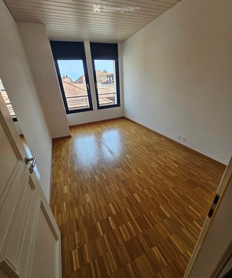 Empty room with wooden flooring, white walls, and large windows offering a view of rooftops.