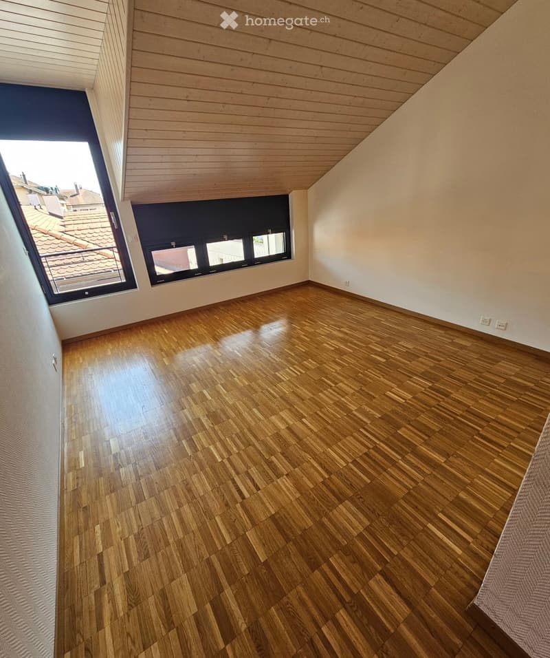 Empty room with wooden flooring, white walls, and large windows offering a view of the roof and surrounding buildings.