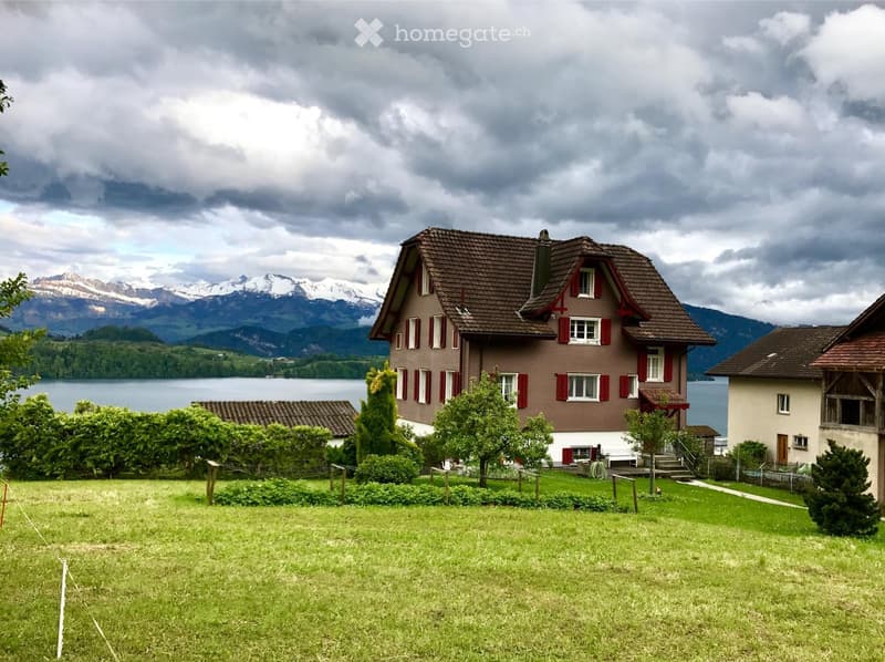 A two-story house with a brown tiled roof and red shutters, situated on a grassy lawn with trees and a view of a lake and mountains in the background.