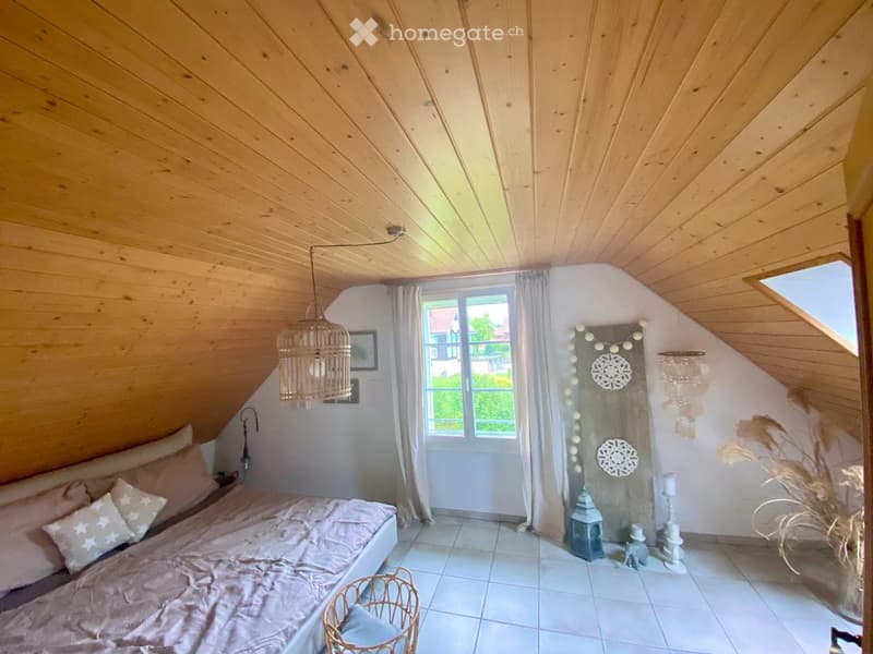 Cozy attic bedroom with wooden slanted ceilings, white tiled floor, large window with white curtains, and a bed with pink bedding and decorative pillows.