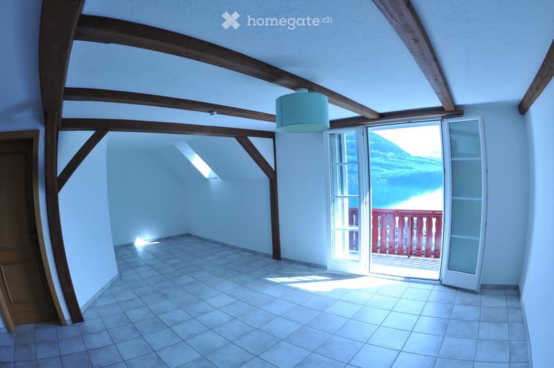 Empty room with exposed wooden beams, tiled floor, and large window opening to a scenic view of a lake and mountains.