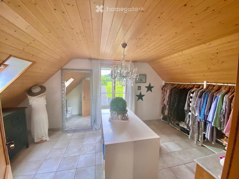 Attic room with wooden slanted ceilings, large mirror, chandelier, clothing rack, small table with plant and cup, and a mannequin with a hat and dress.