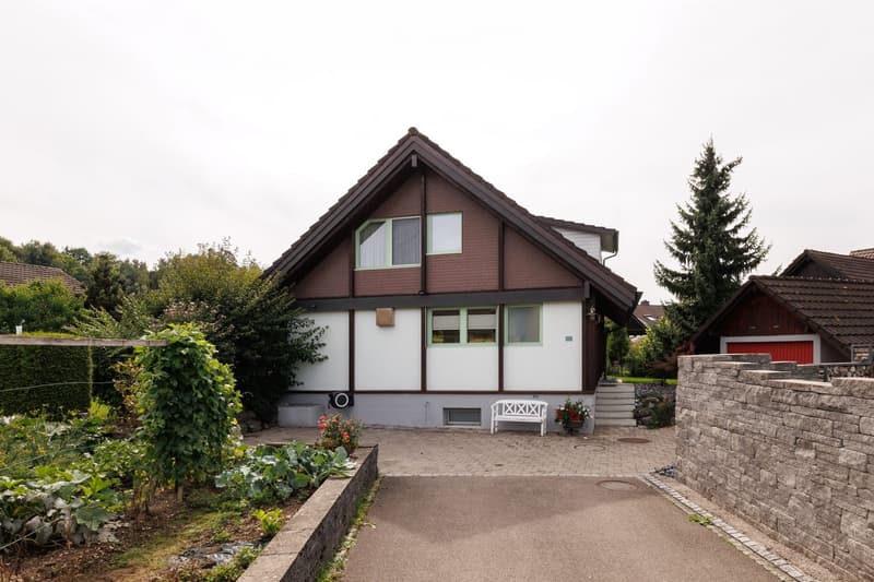 Two-story house with a pitched roof, white and brown exterior, multiple windows, and a small garden area with a bench and plants.