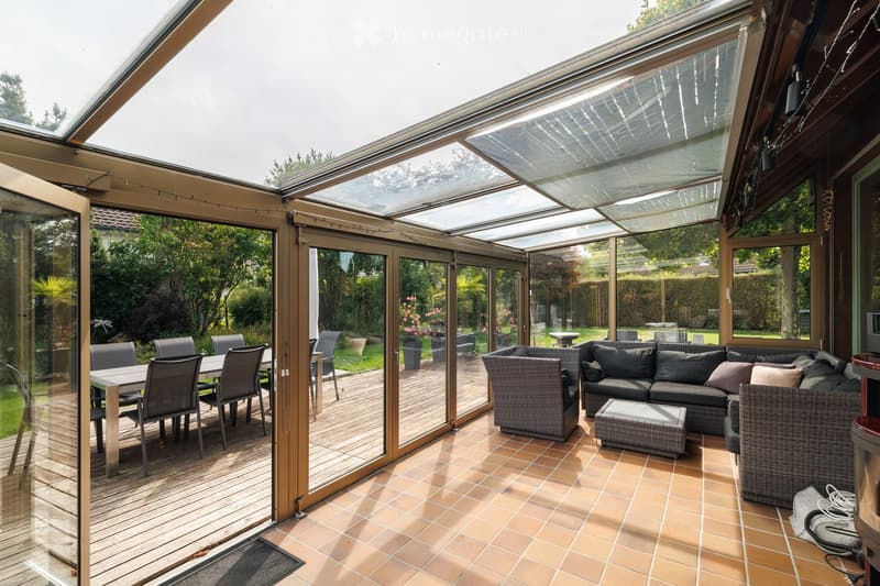 Spacious, well-lit conservatory with tiled flooring, large glass panels, and a comfortable seating area featuring a sofa, ottoman, and wicker chairs. A dining area with a table and chairs is visible through the glass doors.