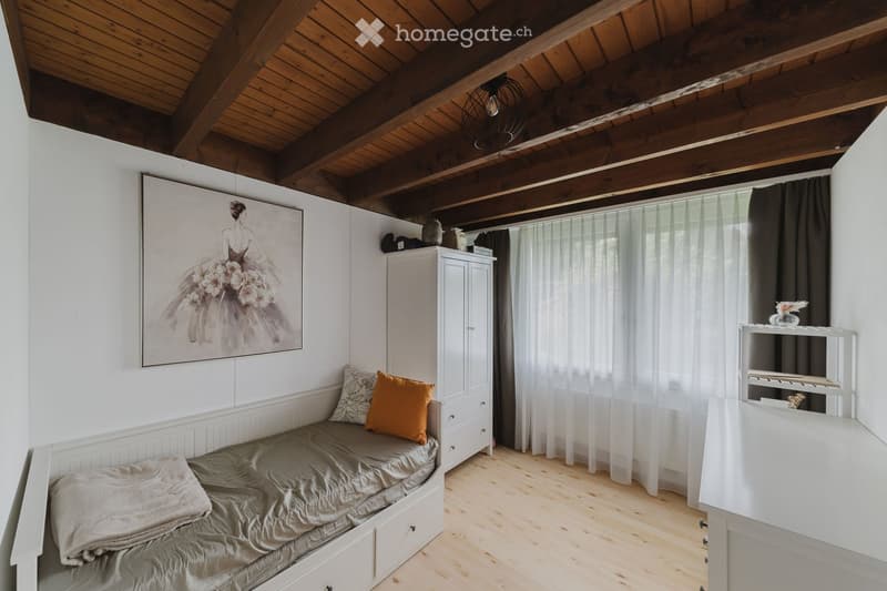 Cozy bedroom with wooden ceiling beams, a daybed with pillows, a white cabinet, and a large window with sheer curtains.