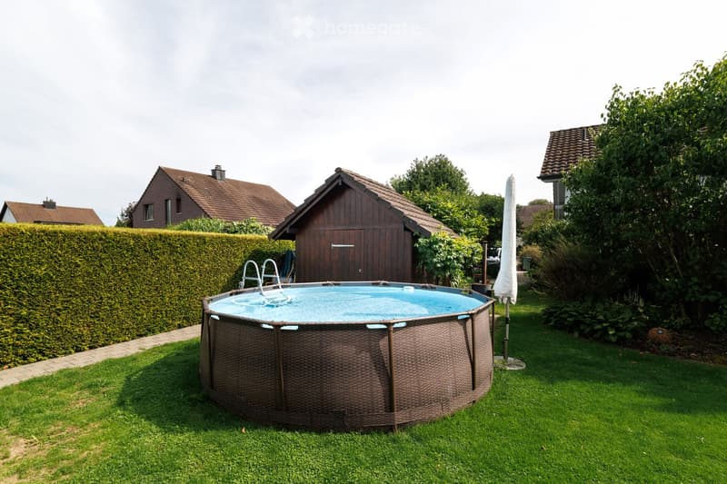 A round, above-ground swimming pool with a wooden frame is situated on a well-maintained lawn, surrounded by greenery and a tall hedge. A wooden shed and several houses are visible in the background.