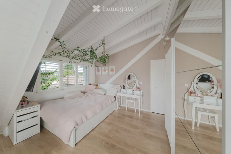 Cozy attic bedroom with slanted white wooden ceilings, light pink bedding, white furniture, and a vanity area with a large round mirror.
