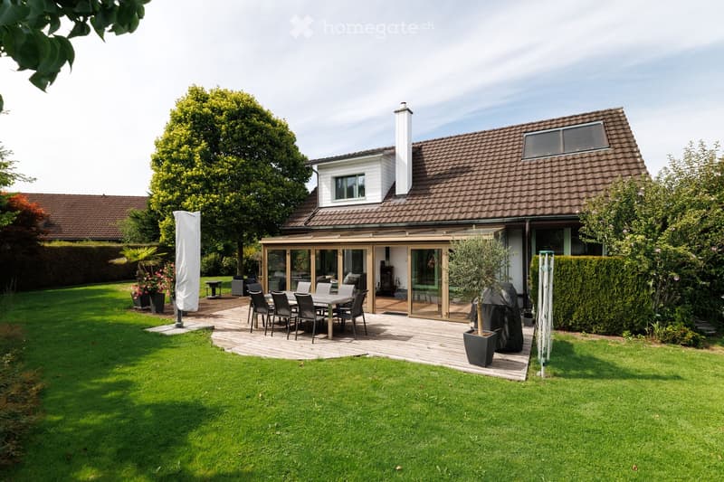 A well-maintained garden with a wooden patio, a dining table with chairs, a large umbrella, and potted plants. The house features a modern design with large windows and a covered outdoor area.