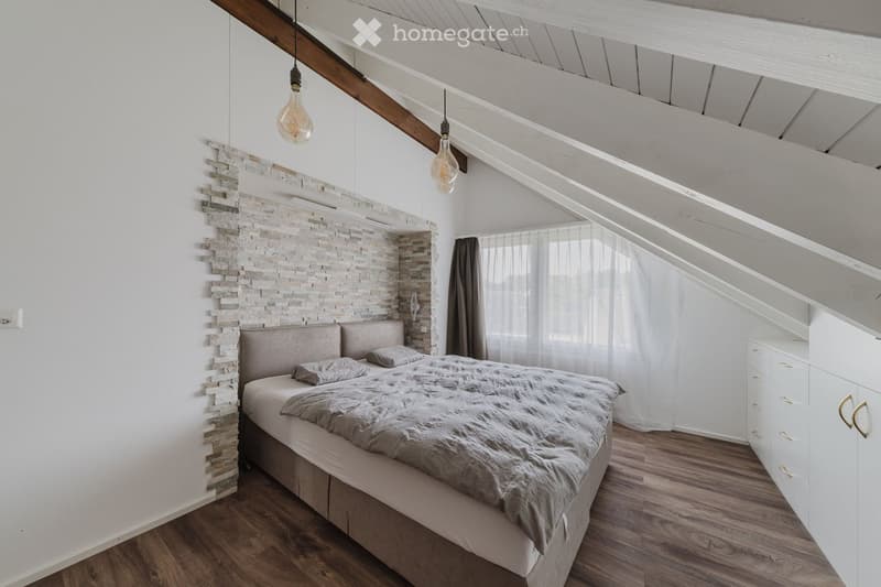 Modern bedroom with wooden flooring, white walls, exposed wooden beams, and a built-in wardrobe. Features a bed with a grey blanket and pillows, a decorative stone accent wall, and large windows with sheer curtains.