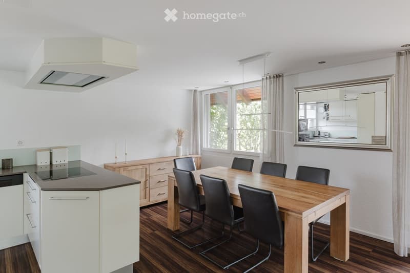 Modern kitchen with a central island, dining table with six chairs, and a large mirror on the wall. The space features wooden flooring, white cabinets, and a sleek design.