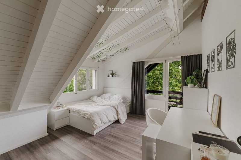 Bright, modern bedroom with a slanted wooden ceiling, large windows, and skylights. Features a white bed, nightstand, desk, chair, and decorative wall art.