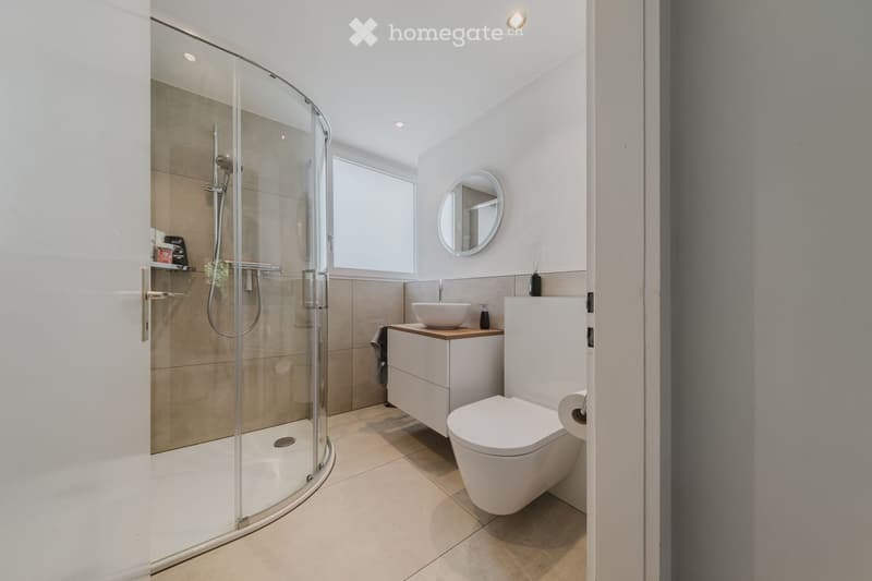 Modern bathroom with a white toilet, sink, and shower enclosure. Features a round mirror, a small shelf, and a window providing natural light.