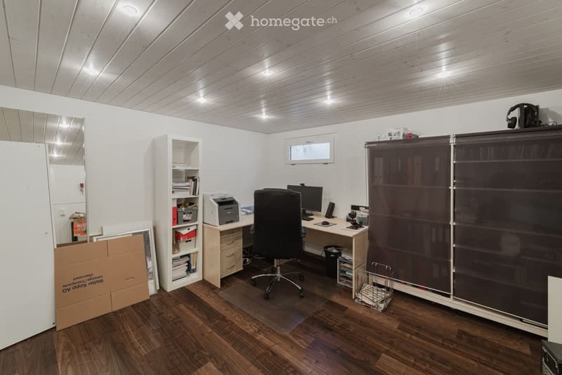 Office space with wooden flooring, white walls, and a slanted wooden ceiling. Includes a desk with a computer, chair, printer, and shelves with books and office supplies. A large mirror and a cardboard box are also visible.