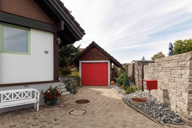 A two-story house with a red garage door, a white bench with flowers, and a stone wall with a mailbox.