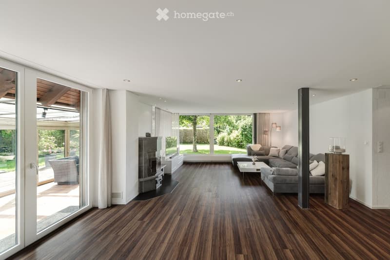Modern living room with hardwood flooring, large windows, fireplace, sectional sofa, coffee table, armchairs, and lamps.