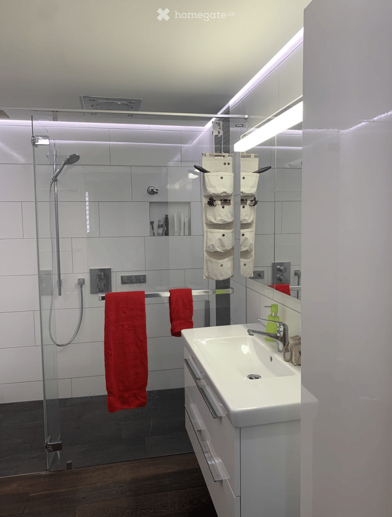 Modern bathroom with white tiles, a glass-enclosed shower, a white sink with a chrome faucet, a large mirror, and a red towel hanging on a rack.