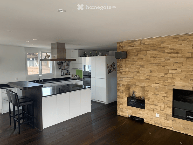 Modern kitchen with white cabinets, dark countertops, a central island with bar stools, and a stone accent wall with a built-in TV.