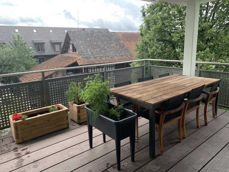 Balcony with wooden decking, metal railing, and outdoor furniture including a table, chairs, and potted plants.