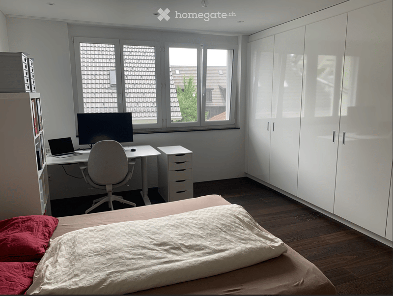 Well-lit bedroom with a bed, desk, chair, laptop, screen, and built-in white cabinets. Large windows offer a view of the roof and surrounding area.