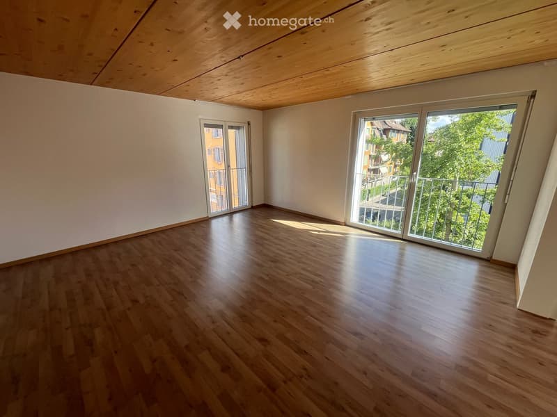 Empty room with wooden flooring, white walls, and large sliding glass doors leading to a balcony with a view of greenery and neighboring buildings.