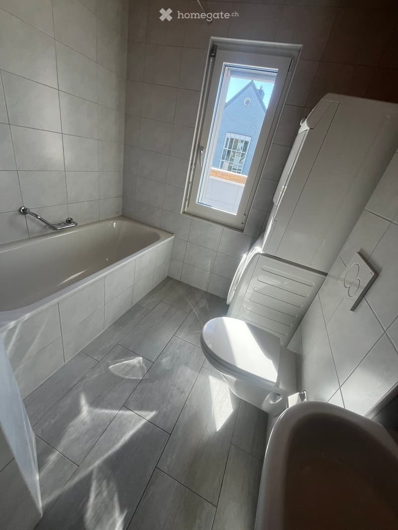 Modern bathroom with a bathtub, toilet, and shower stall. Grey tiled walls and floor, large window providing natural light.