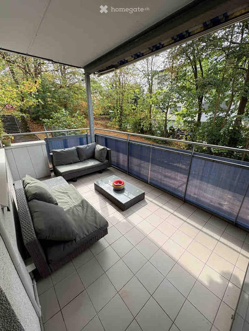 Modern balcony with tiled floor, glass railing, and outdoor seating including a sofa and coffee table.