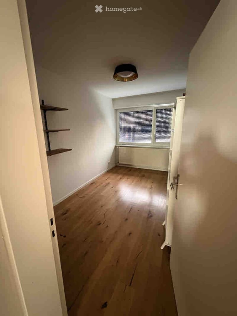 Empty room with wooden flooring, white walls, and a window with blinds. There is a radiator below the window and a small shelf unit on the left side.