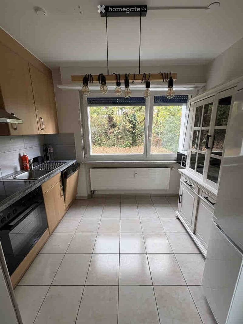 Bright kitchen with white tiled floor, wooden cabinets, sink, oven, radiator, and large windows overlooking a green area.