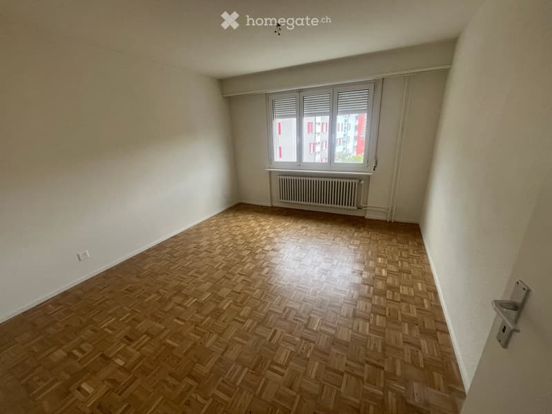Empty room with wooden herringbone flooring, white walls, and a large window with blinds. A radiator is positioned below the window.
