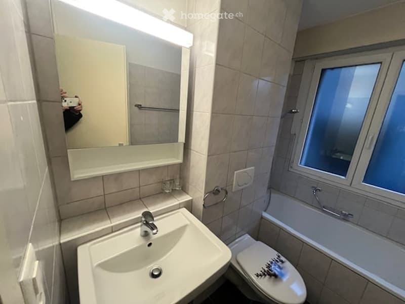 Modern bathroom with a white sink, mirror, toilet, and bathtub. The walls are tiled in beige, and the space is well-lit with a built-in light above the mirror.