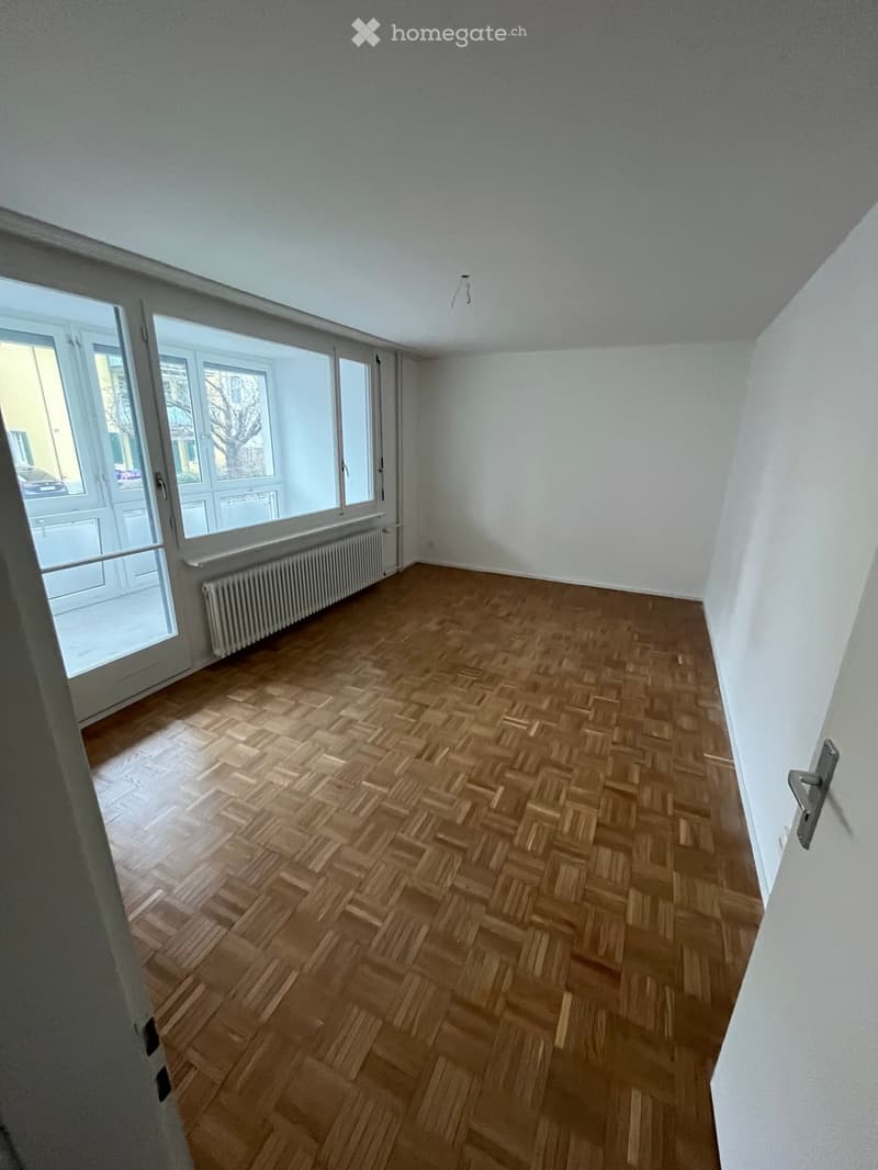 Empty room with wooden herringbone flooring, white walls, large windows, and a radiator under the windows.