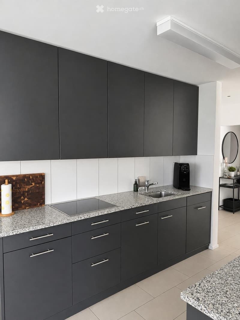 Modern kitchen with dark grey cabinets, granite countertops, built-in electric stovetop, sink, coffee maker, and small shelf with decorative items.