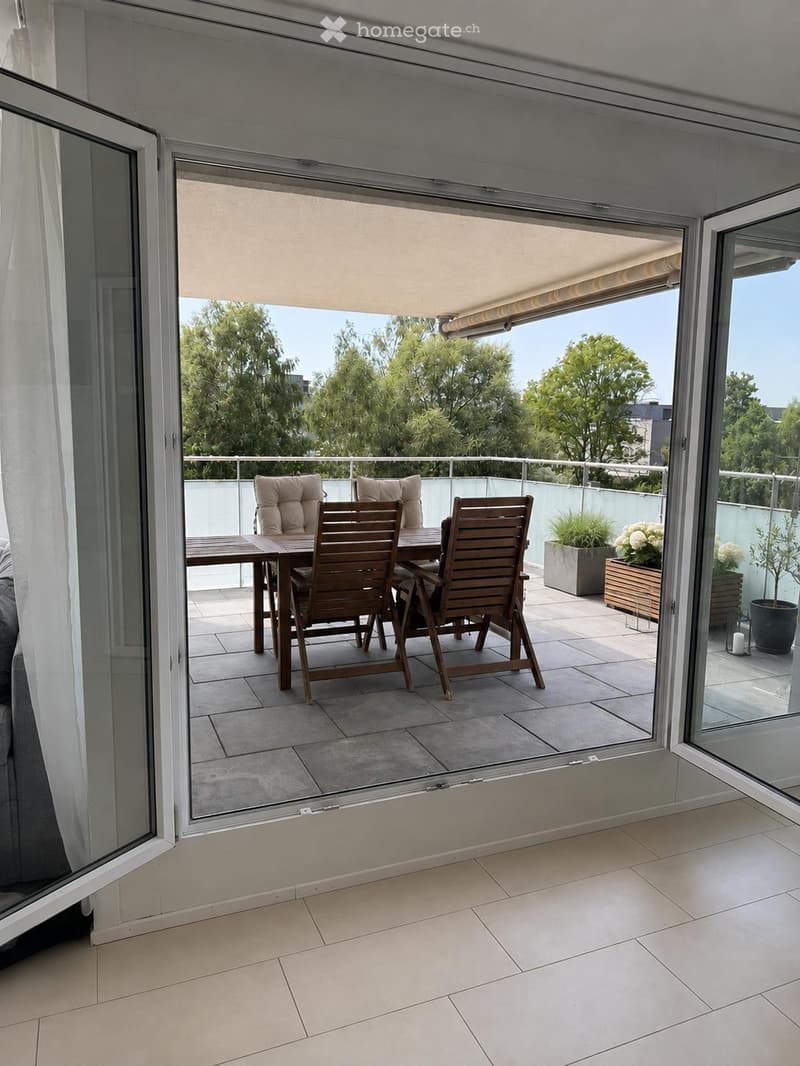 Balcony with a wooden table, chairs, and potted plants, featuring a retractable awning and a view of greenery.