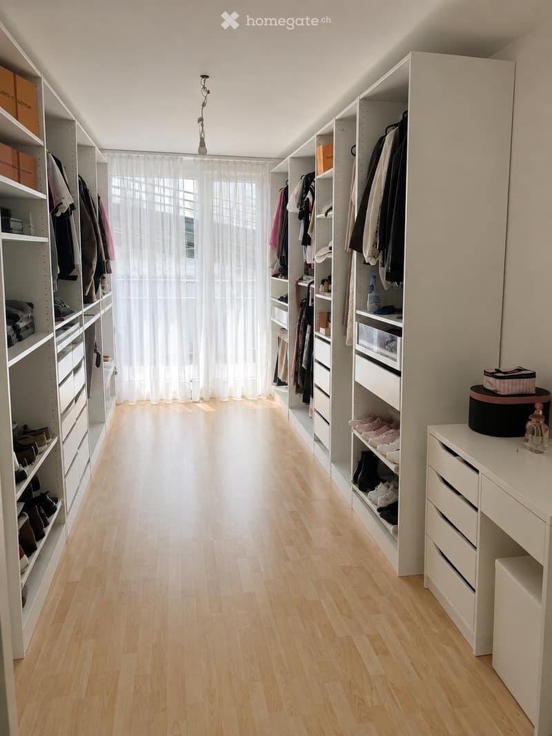 A well-organized walk-in closet with built-in white wardrobes, shelves, and drawers. The space features light wooden flooring, large windows with white curtains, and various clothing and shoes neatly arranged.