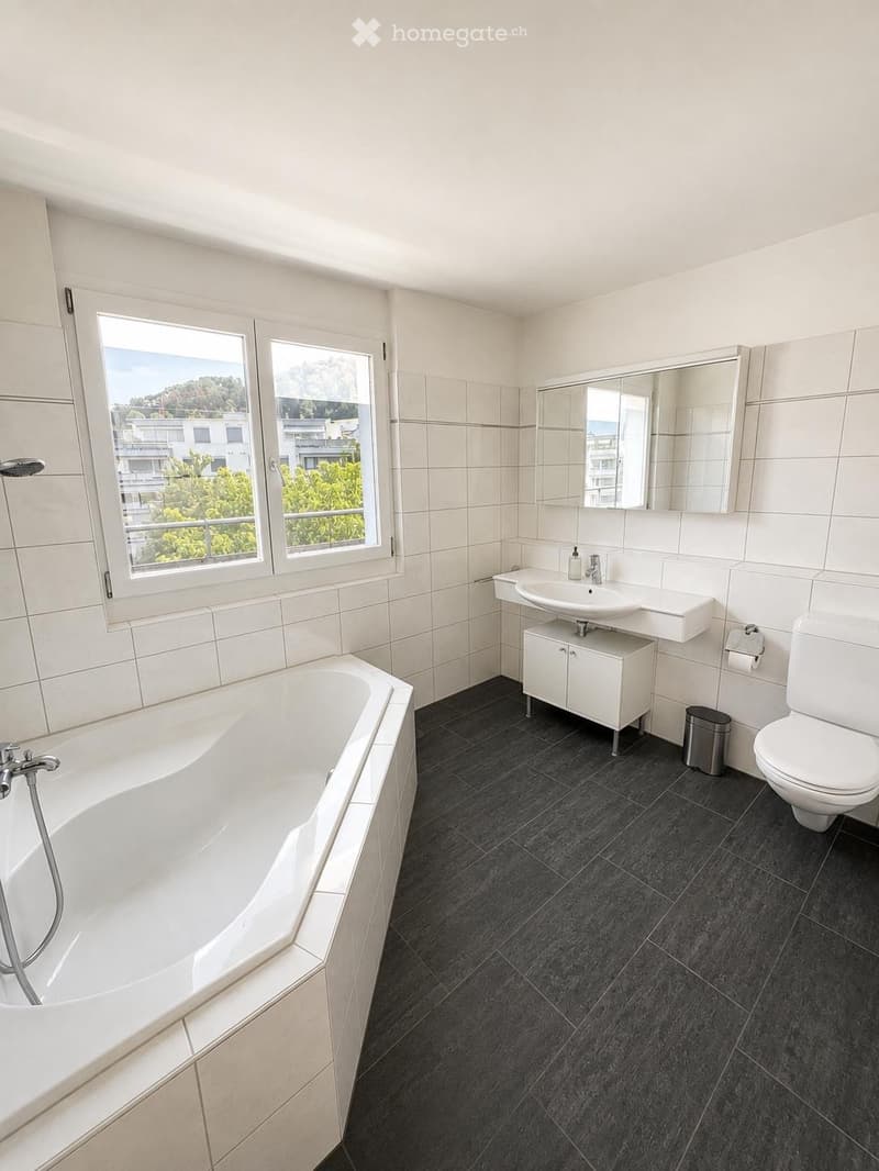 Modern bathroom with white tiles, a large bathtub, a sink with a cabinet, a toilet, and a mirrored cabinet. The room has large windows offering a view of the outside.