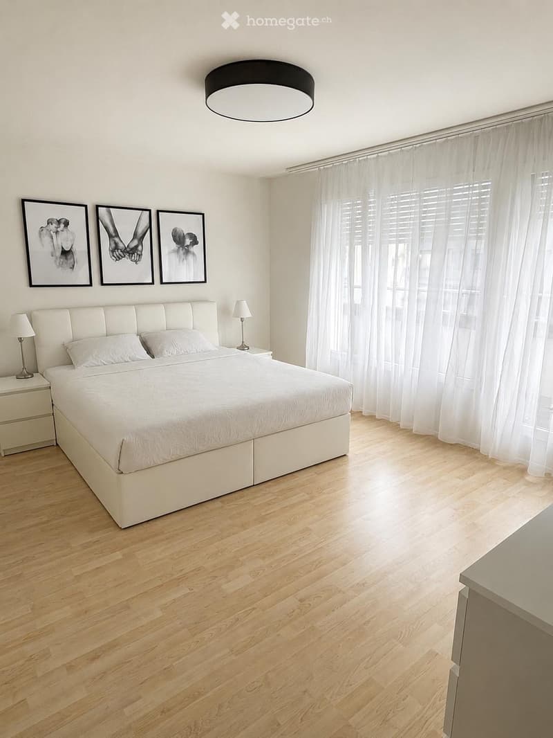 Modern bedroom with a large white bed, nightstands, lamps, and artwork on the wall. The room features wooden flooring and large windows with sheer curtains.