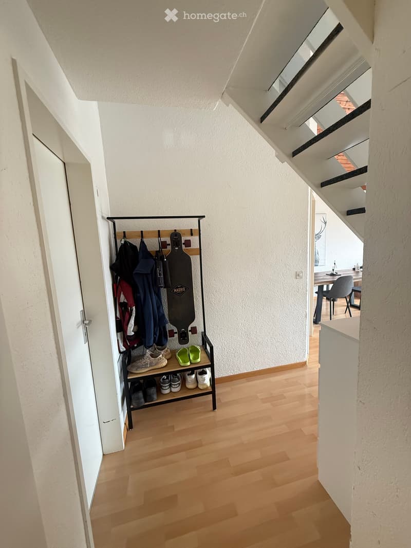 A narrow hallway with wooden flooring, white walls, and a coat rack holding jackets, shoes, and a skateboard. The hallway leads to a dining area with a table and chairs.